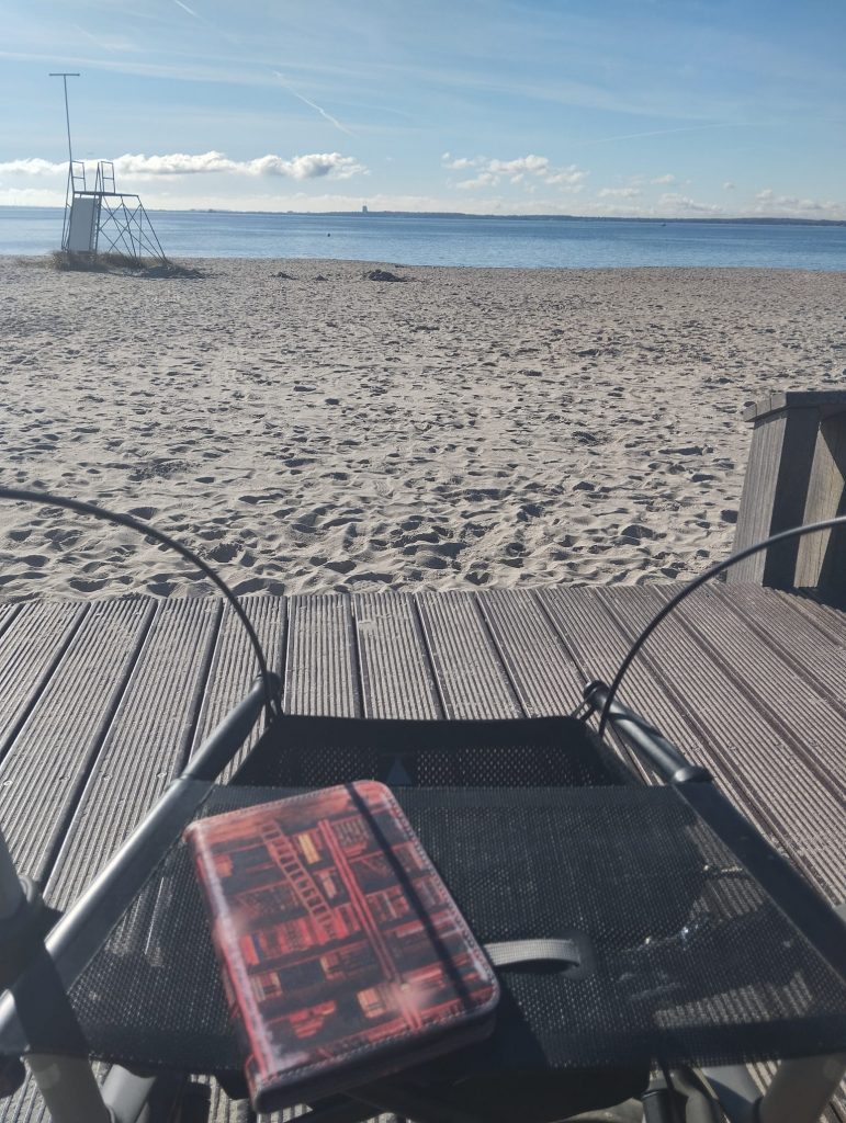 vorne ein Rollator, darauf ein Ebook, dahinter Strand und ganz hinten das Meer