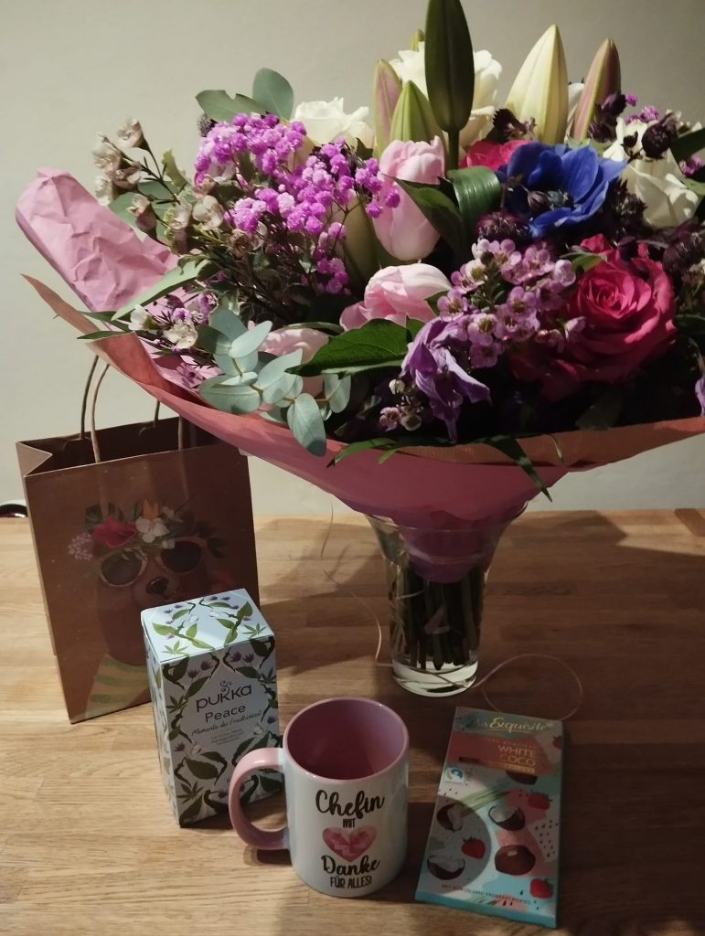 Tisch mit Blumenstrauß, Geschenktüte, Tee, Tasse und Schokolade